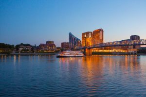 City skyline at dusk along a wide river with a lit bridge and a white boat reflecting warm lights on the water