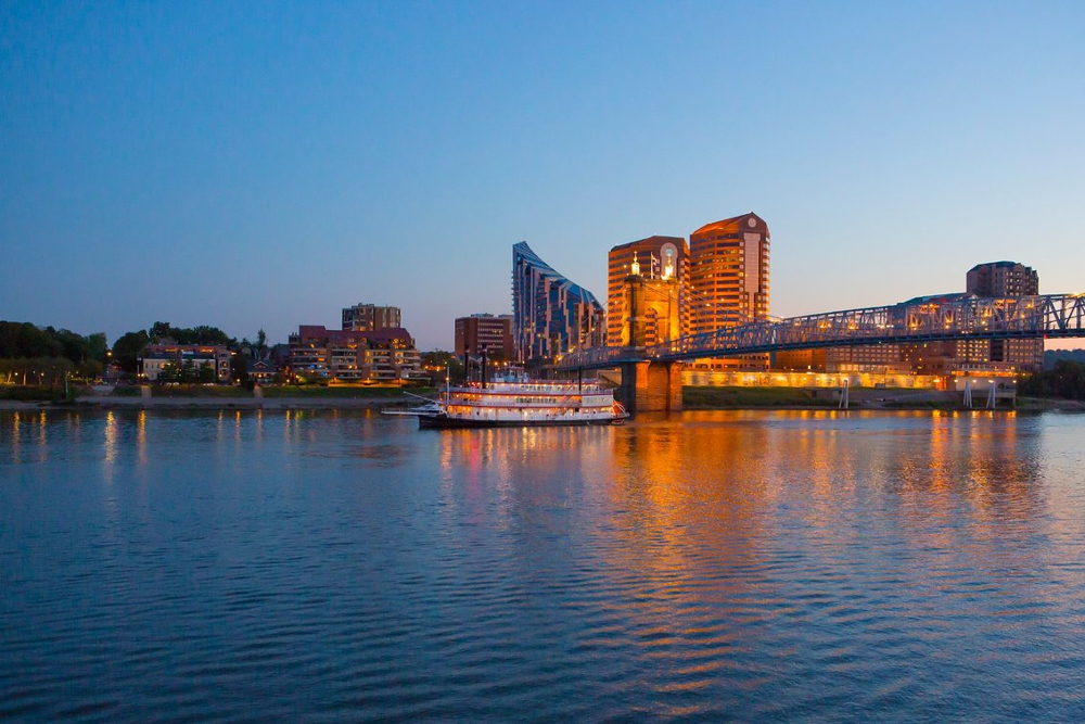 Covington-Kentucky | Route 17 Express City skyline at dusk along a wide river with a lit bridge and a white boat reflecting warm lights on the water