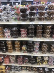 Grocery store shelf filled with clear plastic containers of assorted candies and nuts on multiple levels, including chocolate-covered treats and gummy candies.