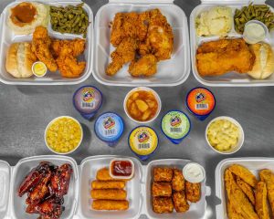 Overhead view of three foam trays with fried chicken, green beans, mashed potatoes, cornbread, and sauce cups on a gray counter.