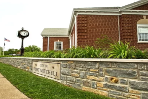Stone wall with a sign reading 'City of Fort Mitchell' in front of a brick municipal building and a tall clock tower; an American flag nearby.