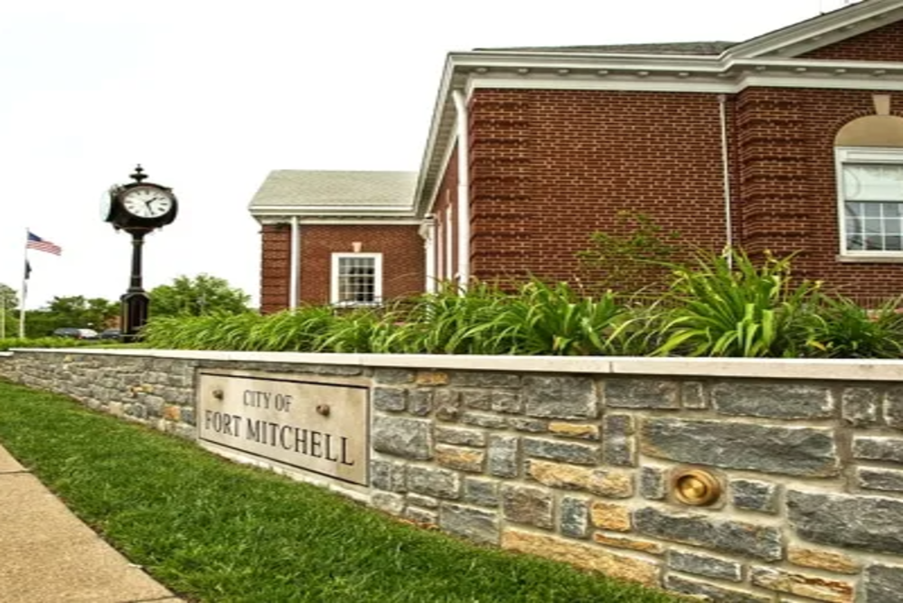 Stone wall with a sign reading 'City of Fort Mitchell' in front of a brick municipal building and a tall clock tower; an American flag nearby.