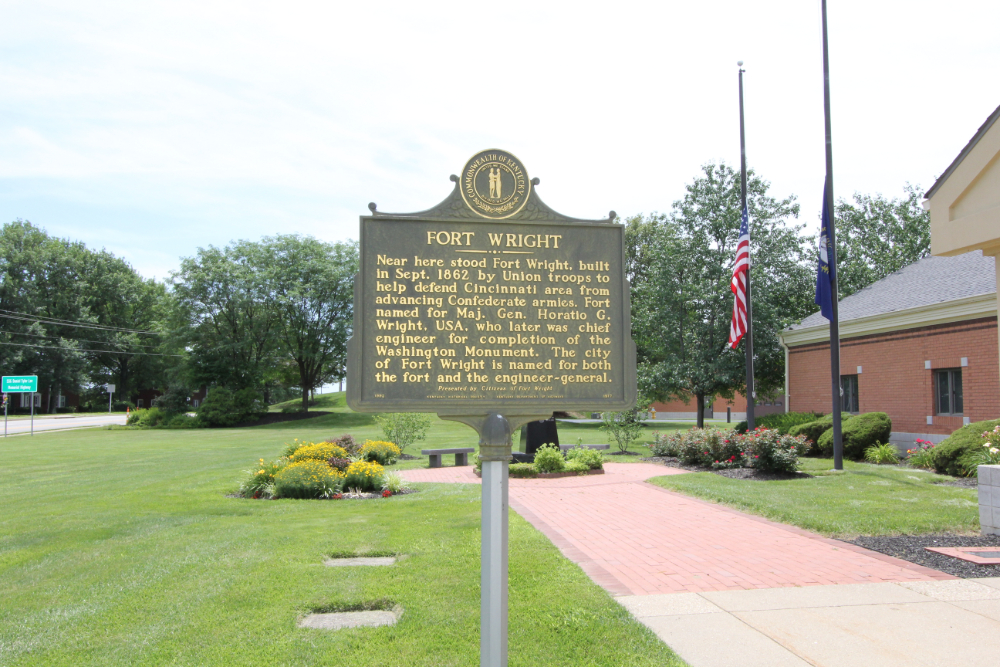 Fort-Wright | Route 17 Express Bronze historical marker labeled 'Fort Wright' on a post, with a flag, brick building, and landscaped lawn in the background on a sunny day.