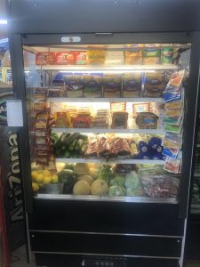 Refrigerated grocery display case filled with assorted cheeses, sausages, and fresh produce on shelves and bottom drawer area