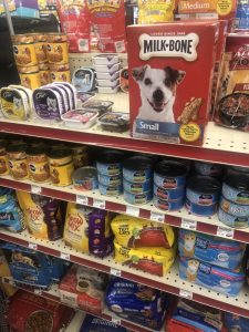 Dog treat shelf featuring a large Milk-Bone box with a smiling dog on the front, surrounded by various canned and bagged pet foods by the checkout area.