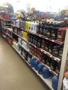 Aisle in a store with metal shelves lined with motor oil and automotive fluids in black, red, and blue jugs on multiple shelves.