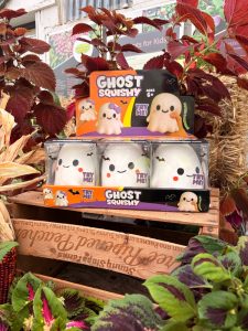 Display of Ghost Squishy toys in ghost-shaped packaging on a wooden crate among autumn red leaves and plants, labeled Ages 6+.