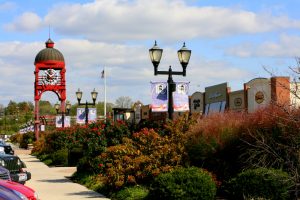 Red clock tower and street lamps lining a suburban shopping district with banners and storefronts under a blue sky.