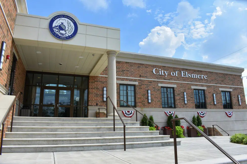 Elsmere | Route 17 Express Front entrance of the City of Elsmere building: brick exterior, glass doors, concrete steps, and patriotic red-white-blue bunting along the railings.